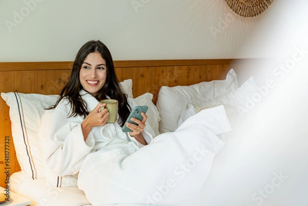 Fototapeta Young woman wearing bathrobe relaxing in bed, drinking coffee and using her smartphone in a hotel room
