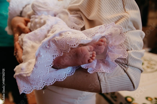 Obraz During the rite of baptism of a child in the church, the godmother holds the child dressed in a magnificent dress in her arms, the child's small legs are in the photographer's frame