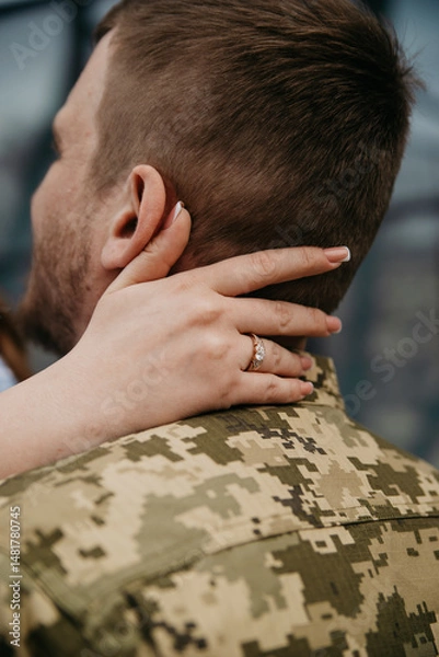 Obraz The groom in the military uniform of the Armed Forces of Ukraine hugs his bride on their wedding day. The bride has a gold wedding ring on her fingers, and the bride's hand hugs the groom's neck