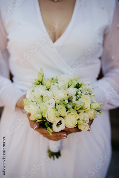 Obraz The bride in a trendy white wedding dress holds a bouquet of white roses wrapped together with a white ribbon in front of her with both hands