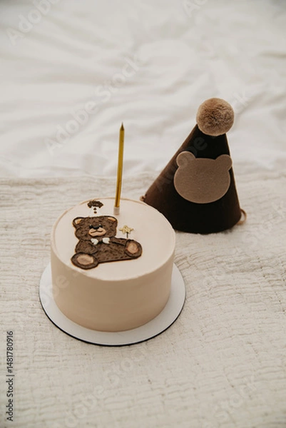Obraz a small cake or cupcake with one candle inserted in it and decorated with a decorative chocolate bear on an isolated background for a birthday celebration and a festive hat on the head next to it