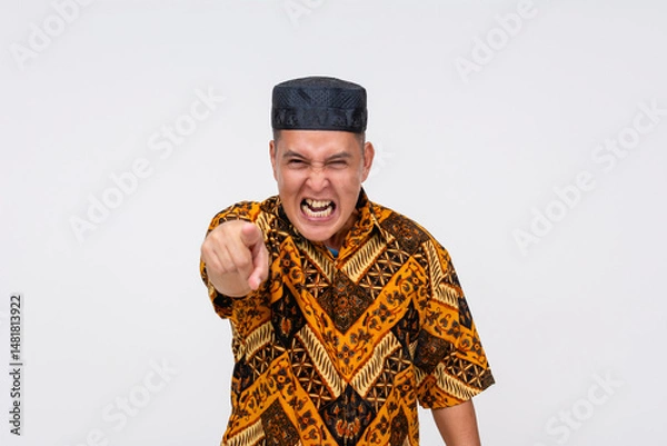 Fototapeta Furious middle-aged Indonesian man shouting while pointing finger aggressively, wearing Batik shirt and black songkok. Isolated on a light background.