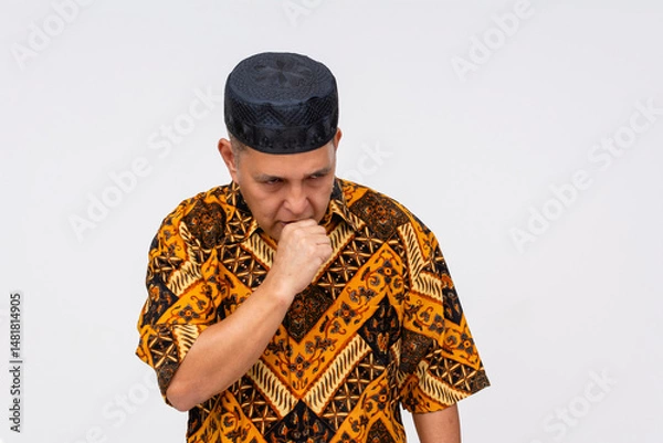Fototapeta Thoughtful middle-aged Indonesian man looking down with hand near mouth, showing concern, dressed in Batik and songkok. Isolated on a light background.