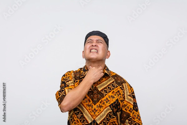 Fototapeta Middle-aged Indonesian man wincing in pain, clutching his throat with discomfort while wearing Batik shirt and songkok. Isolated on a light background.