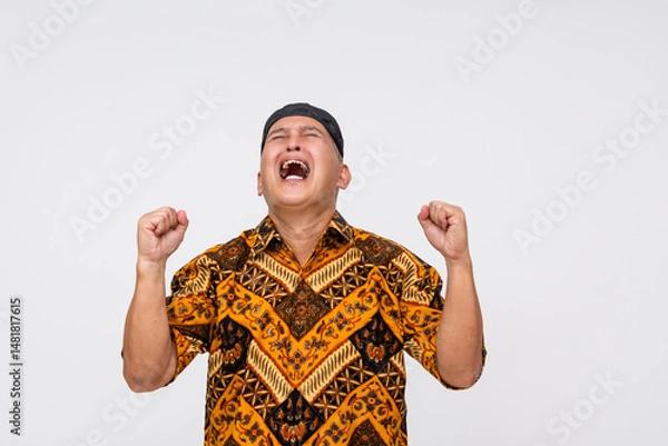 Fototapeta Frustrated Indonesian man shouting upward with clenched fists in emotional outburst, dressed in traditional Batik shirt and black cap. Isolated on a light background.