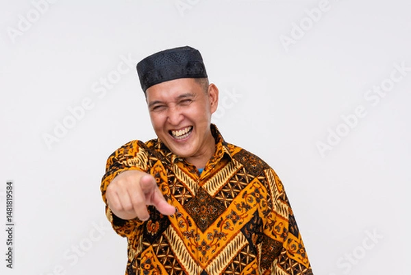 Fototapeta Middle-aged Indonesian man bursting into laughter while pointing directly ahead, dressed in Batik shirt and traditional songkok. Isolated on a light background.