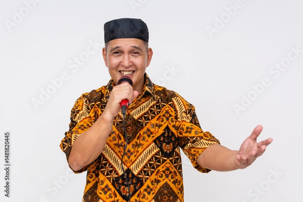 Fototapeta Smiling Indonesian man speaking into microphone while gesturing openly, wearing vibrant Batik shirt and black songkok. Isolated on a light background.