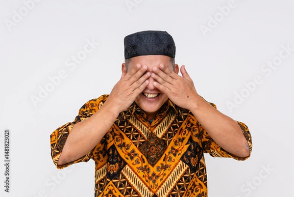 Fototapeta Smiling Indonesian man playfully covering his eyes with both hands, showing cheerful mood in Batik shirt and headwear. Isolated on a light background.