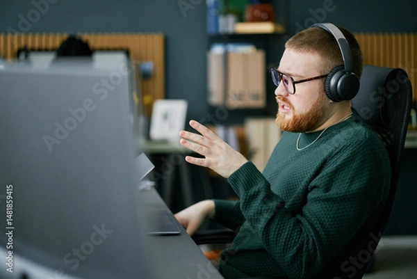 Fototapeta Man wearing glasses and headphones, working on computer at desk in modern office. Bookshelf and wall decor visible in background adding to atmosphere