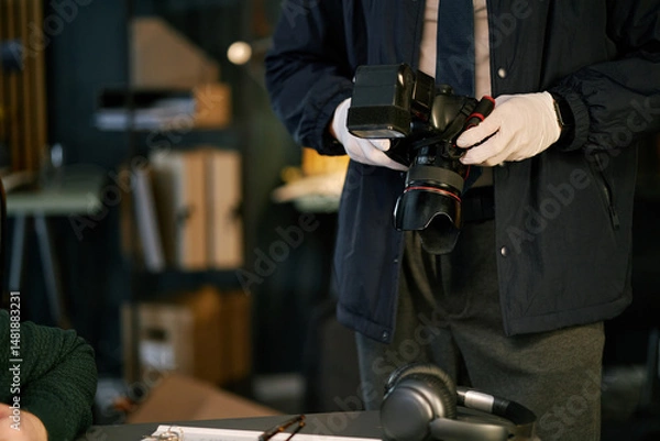 Fototapeta Person holding professional camera while preparing for photography indoors, with bookshelf visible in background creating cozy atmosphere. Environment features dim lighting and warm tones