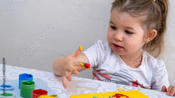 Fototapeta Happy smiling little girl enjoying creative finger painting activity, learning colors and shapes, improving fine motor skills through fun art and early childhood education.