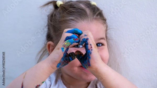 Fototapeta Smiling Little Girl Making Heart Shape with Colorful Painted Hands, Playful and Creative Expression During Fun Finger Painting Activity, Early Childhood Art Concept