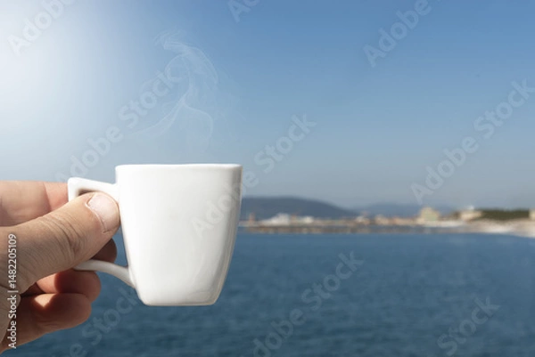 Fototapeta Man holds a cup of coffee in front of the sea