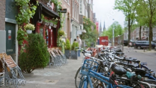 Fototapeta Blurred bicycles line a charming amsterdam street with vibrant shops and greenery, capturing the essence of urban life in the netherlands.