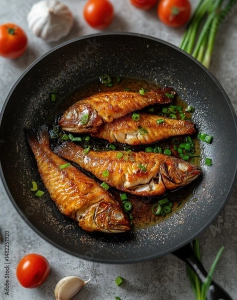 Fototapeta top view of golden fried fish in a black pan with oil and green onions next to it on a grey concrete table.