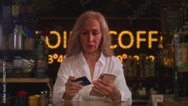 Obraz An old woman uses a credit card to shop on a smartphone. Senior female makes a purchase in an online store. Order online, enter the card number.