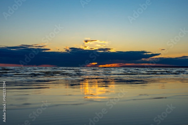 Fototapeta Colorful sunset on the sea beach. Beautiful reflections of the sun on the water, calm waves, soft sandy beach against the cloudy sky.