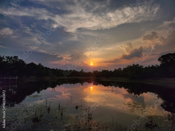 Fototapeta Stunning sunset over a tranquil lake with vivid reflections and dramatic clouds. Ideal for backgrounds, nature themes, travel, and inspirational content. Perfect peaceful nature scene.