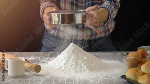 Fototapeta Artisan bread dough preparation. Use sieve or colander to sift flour into pile of flour for handmade bread with bakery equipment tool, black background. professional baker process at work.
