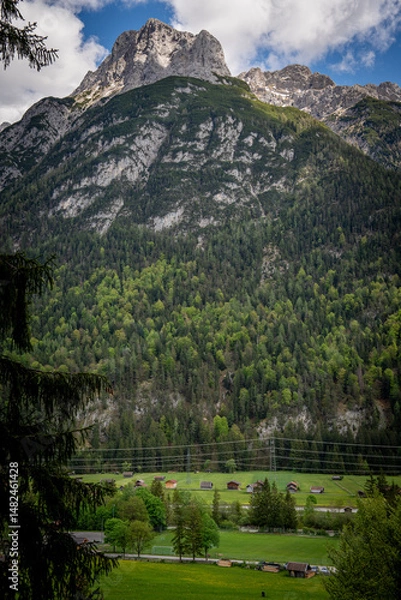 Fototapeta Imposing Linderspitze Mountain In Karwendel Range: Towering Rocky Peak Rising Above Dense Forests And Charming Alpine Meadow With Scattered Houses Under A Dramatic Sky In Tyrol, Austria