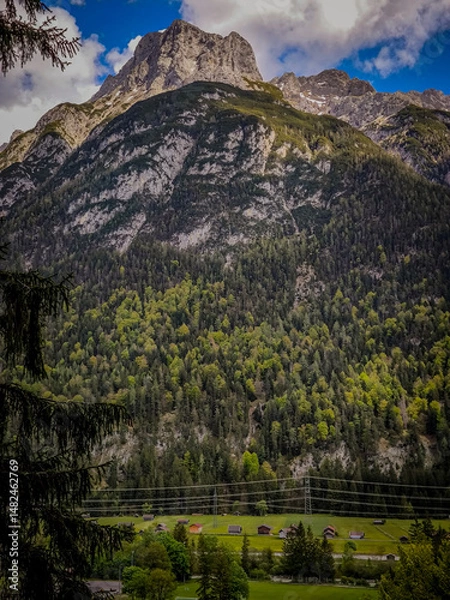 Fototapeta Imposing Linderspitze Mountain In Karwendel Range: Towering Rocky Peak Rising Above Dense Forests And Charming Alpine Meadow With Scattered Houses Under A Dramatic Sky In Tyrol, Austria