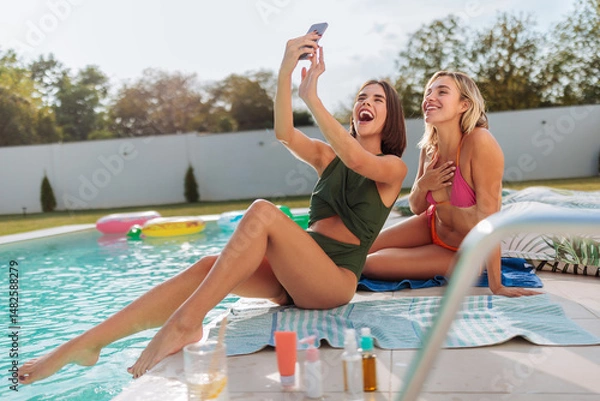 Fototapeta Two young women taking photo of themselves by the poolside