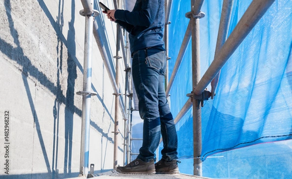 Obraz Engineer standing inside scaffolding with scaffold netting. Side view of female bylaw inspector or project manager on construction site taking a picture or documenting notes. Selective focus.