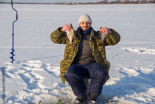 Fototapeta A male fisherman sits on the ice of a lake and holds a fish he caught in his hands. He looks at the camera.