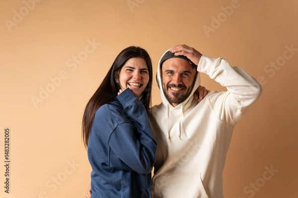 Fototapeta Happy couple laughing and embracing, wearing hoodies, on beige background