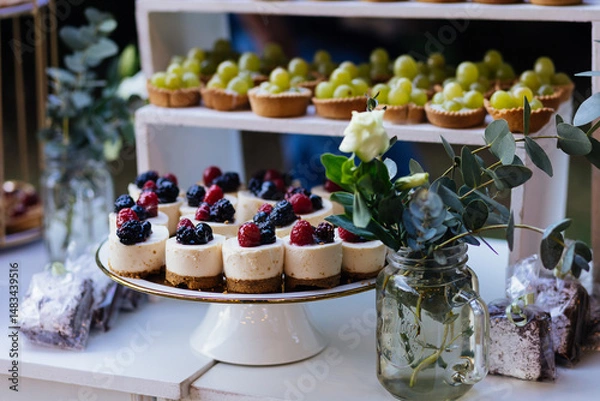 Fototapeta A beautifully arranged dessert table featuring mini berry cheesecakes, green grape tarts, and floral decor. The clean, natural style makes it ideal for events, catering, and gourmet branding. 