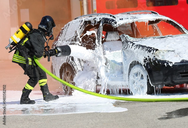 Fototapeta Firefighter with helmet and oxygen cylinder while operating   lance with   foam to extinguish   car fire after   road accident