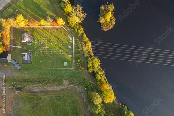 Fototapeta Small electrical substation near a river, surrounded by nature, with visible power lines and infrastructure