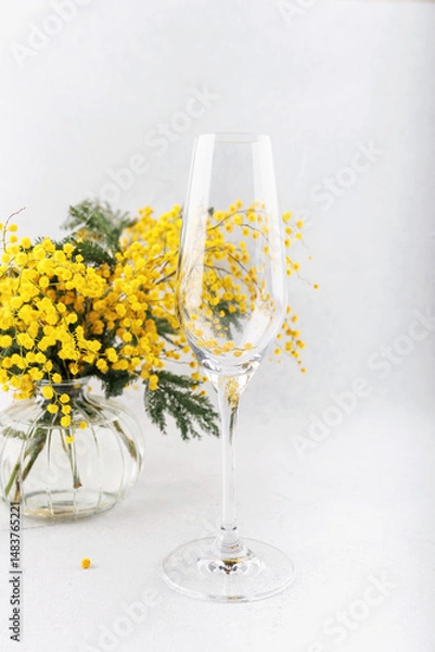 Fototapeta A close-up of a champagne flute reflecting yellow mimosa flowers from a glass vase, creating a delicate and artistic floral composition.