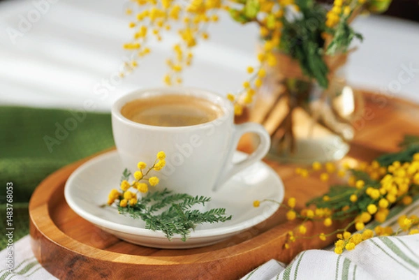Fototapeta A cozy spring morning scene with a white coffee cup on a wooden tray, decorated with fresh mimosa flowers, exuding warmth and charm.