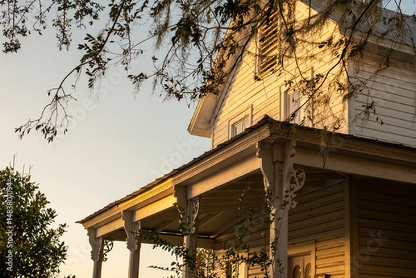 Fototapeta A vintage roof of a farmhouse with multiple stories, glass windows, porch posts, decorative face brackets, and cedar shingles. There are tall trees around the building. The sun is setting.