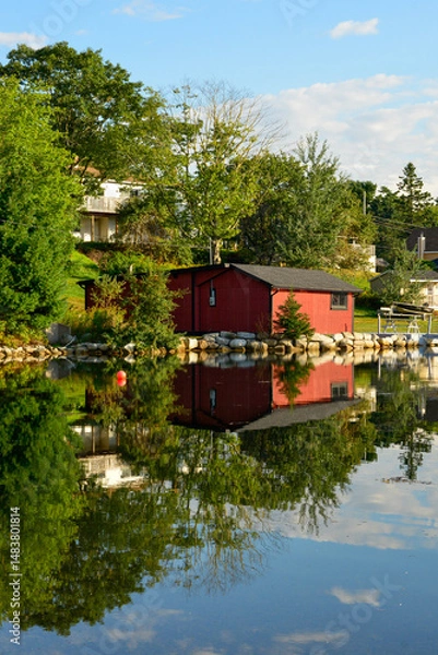 Fototapeta A small red wooden boathouse on the side of a river. The water is calm and still. The red building and lush green trees are reflecting or mirroring in the ocean. The shore is reflected in the sea.