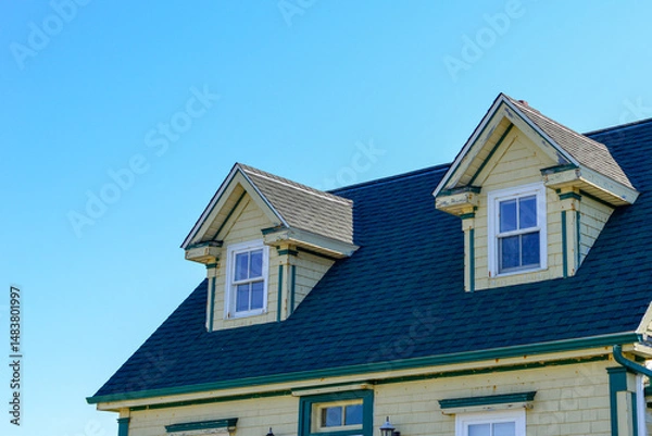 Fototapeta The top exterior floor of a yellow colored Cape Cod house with green and white trim. The building has two peaked dormer windows. The roof is covered in black asphalt shingles. The sky is bright blue.