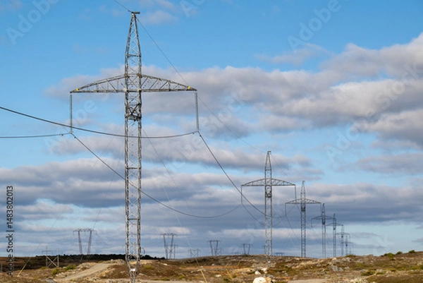 Fototapeta A metal electric utility pole with electrical wires, transmission lines, conductors, and distribution wires for high voltage electricity. The pole has a blue sky in the background with white clouds.