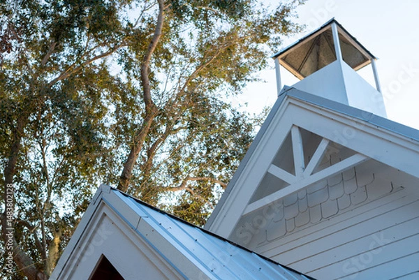Fototapeta The end of a peaked roof has a decorative bracket. The wood, metal, and small cedar shakes are white. A square, vented cupola sits on top of the church-like building. The roof has grey metal panels.