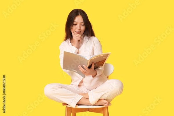 Fototapeta Beautiful young Asian woman in pajamas with magazine sitting on chair against yellow background