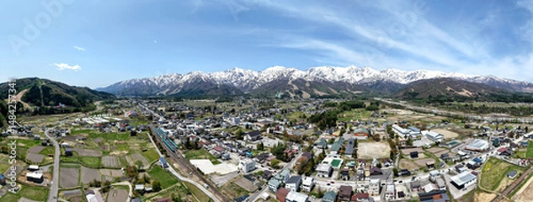 Fototapeta Panoramic view of a peaceful rural town nestled in a valley with snow-capped mountains towering in the background under a clear blue sky at Hakuba Mountain Nagano, Japan.
