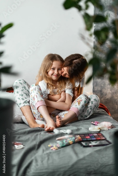 Fototapeta Laughing mother kisses her daughter while spending time together at home. Pajamas, makeup items, and soft pillows create cozy and joyful atmosphere of family bonding in relaxed morning scene.