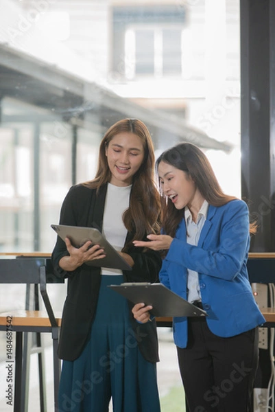 Fototapeta Two  Asian businesswomen holding iPads, holding graph papers, standing in front of the glass, talking and consulting with each other.