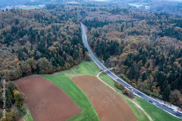 Fototapeta Highway Construction Site Stretching Through Farmland, Forest, and Distant Village Captured by Drone Under Overcast Sky
