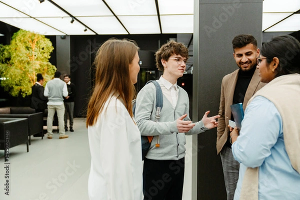 Fototapeta A multinational group of young people in business attire is standing indoors in a modern office lobby during an internship program, with a young White man holding notebooks and gesturing.