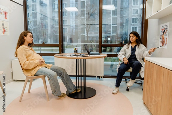 Fototapeta An Indian female doctor is sitting on a chair and pointing with a stick at a poster of a pregnant woman on a wall while looking at a White pregnant woman who is sitting at a table in front of her