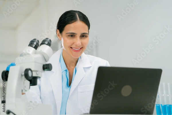 Fototapeta scientist and researcher in laboratory setting smiles while working on laptop, with microscope and test tubes nearby, showcasing chemistry research. scientist researcher chemistry laboratory