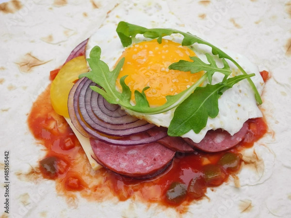 Fototapeta Unwrapped thin pita bread with fried egg with arugula leaf on slices of sausage, cheese, rings of red onion and yellow paprika for grill. Variant of healthy summer meal and hearty breakfast.