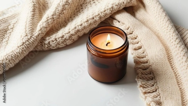 Obraz A cozy lit candle in a brown glass jar sits beside a knitted beige blanket on a white surface, creating a warm and relaxing atmosphere.