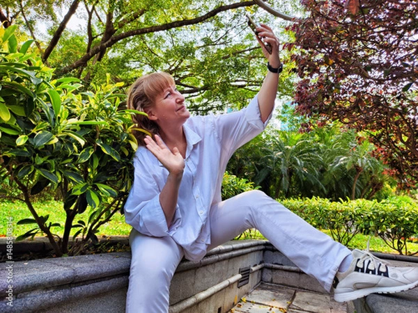 Fototapeta Middle-aged blonde woman enjoys a selfie while exploring a lush park during her travel adventure